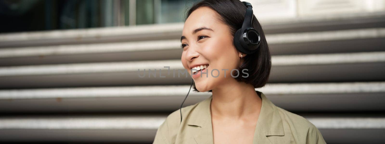 Smiling asian girl, laughing, listening music in headphones, sitting outdoors. Uni student enjoying free time by Benzoix
