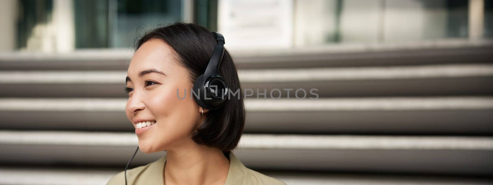 Smiling asian girl, laughing, listening music in headphones, sitting outdoors. Uni student enjoying free time.