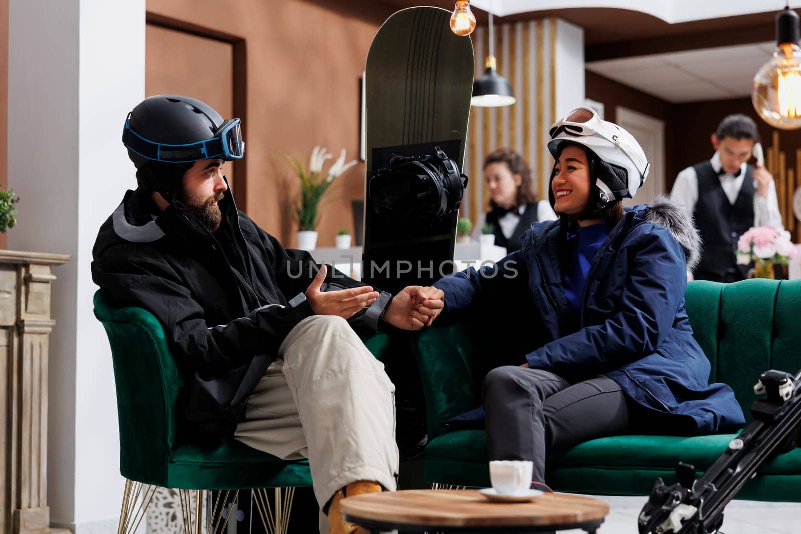 Boyfriend and girlfriend in possession of wintersports equipment engaged in dialogue about plans for skiing and snowboarding. Enthusiastic couple warmly dressed for snow activities seated in lobby.