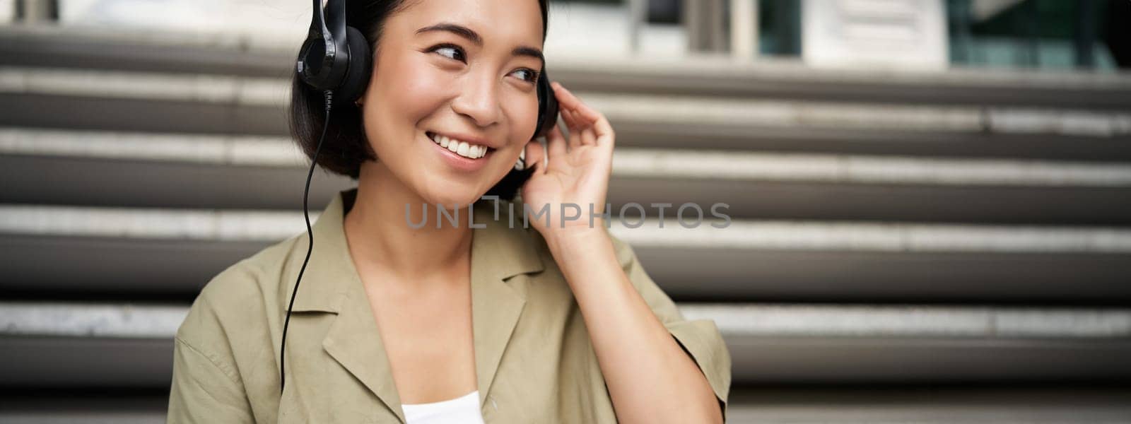 Portrait of beautiful girl in earphones, listening music, smiling and walking in city centre by Benzoix