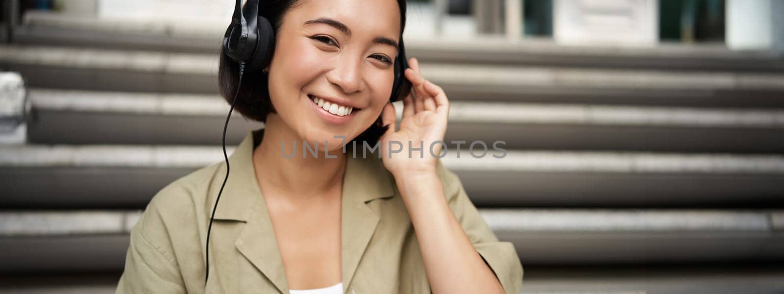 Happy asian girl listens to music in headphones, smiles, sits outdoors on stairs by Benzoix