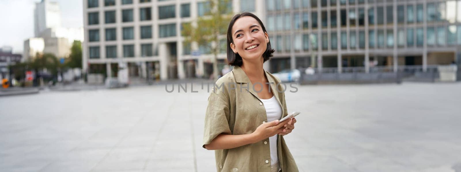 Portrait of asian woman standing on street, city square and holding mobile phone. Girl with smartphone walking outdoors by Benzoix