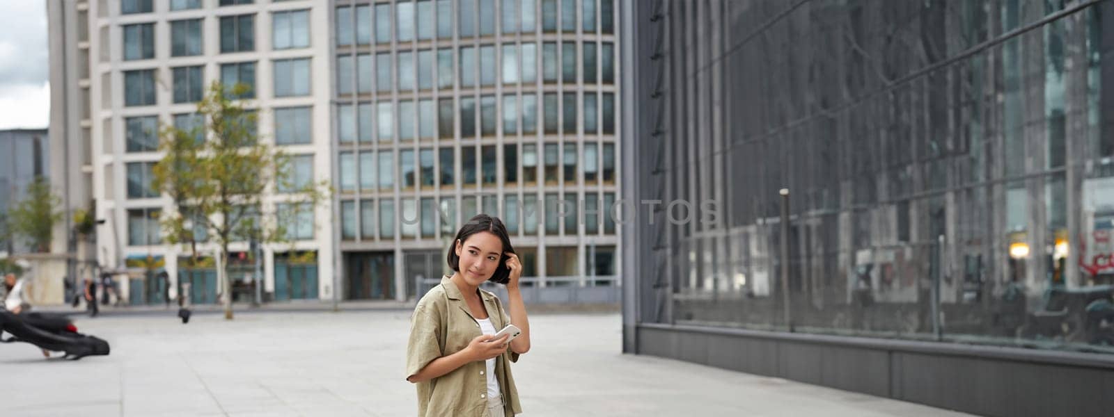 Portrait of asian woman walking in city. Street style shot of girl with smartphone, posing outdoors on street by Benzoix