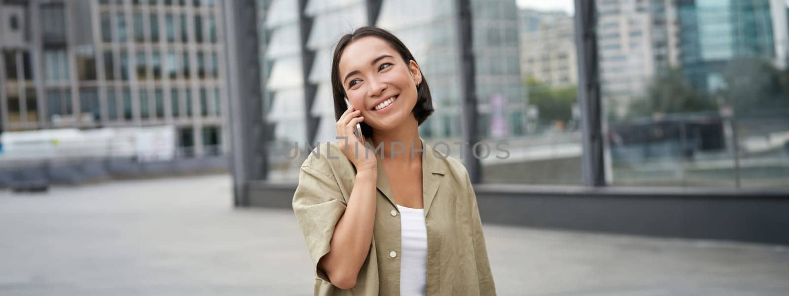 Smiling young korean girl talking on mobile phone and walking in city. Happy woman posing on street with smartphone.
