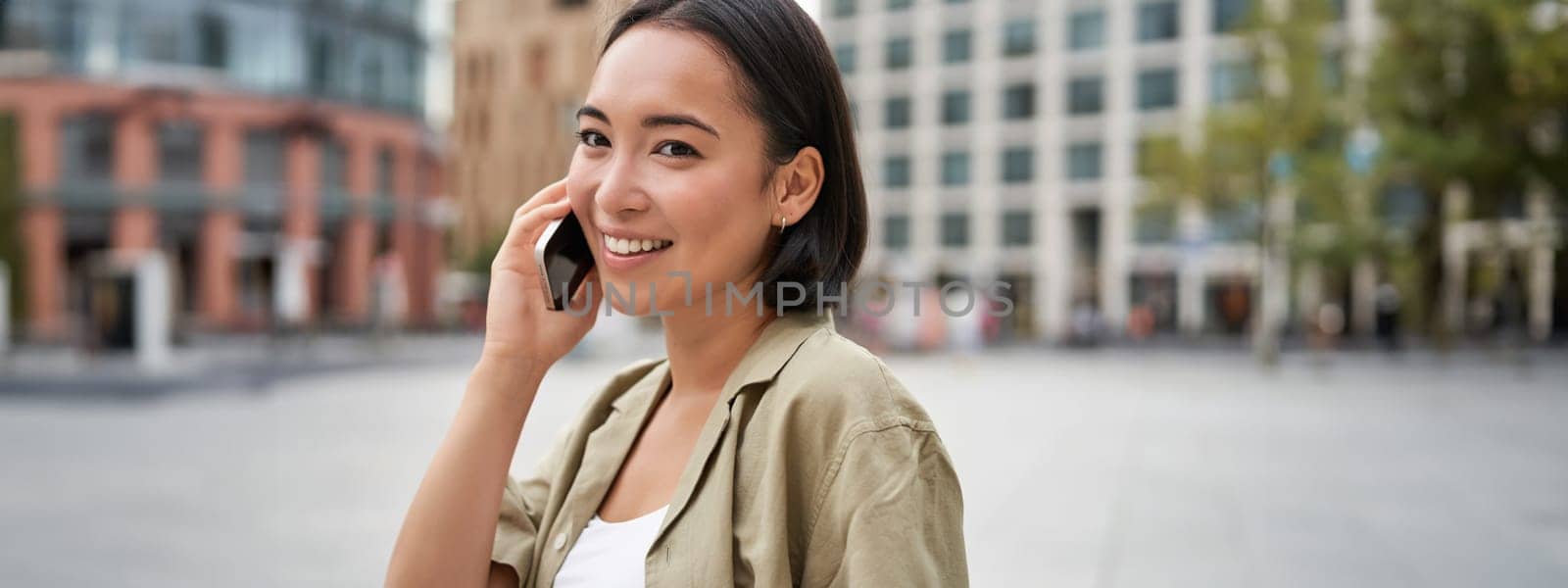 Cellular connection. Young asian woman makes a telephone call, talking on mobile smartphone and walking on street.