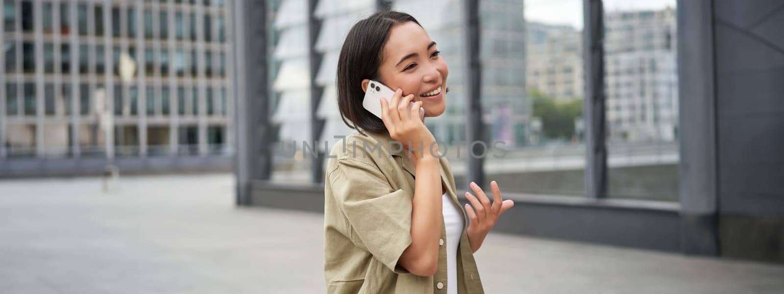 Cellular connection. Young asian woman makes a telephone call, talking on mobile smartphone and walking on street by Benzoix