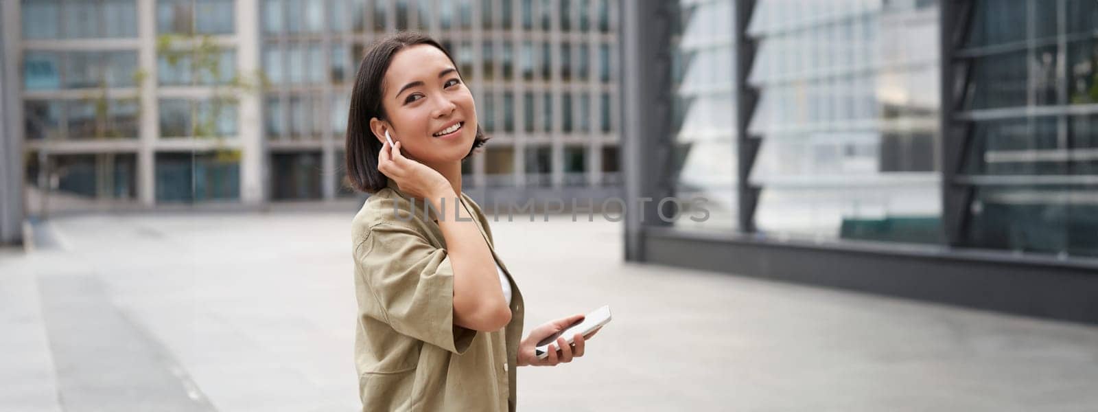 Modern people. Young woman walks on street of city and listens music in wireless headphones, holds smartphone.