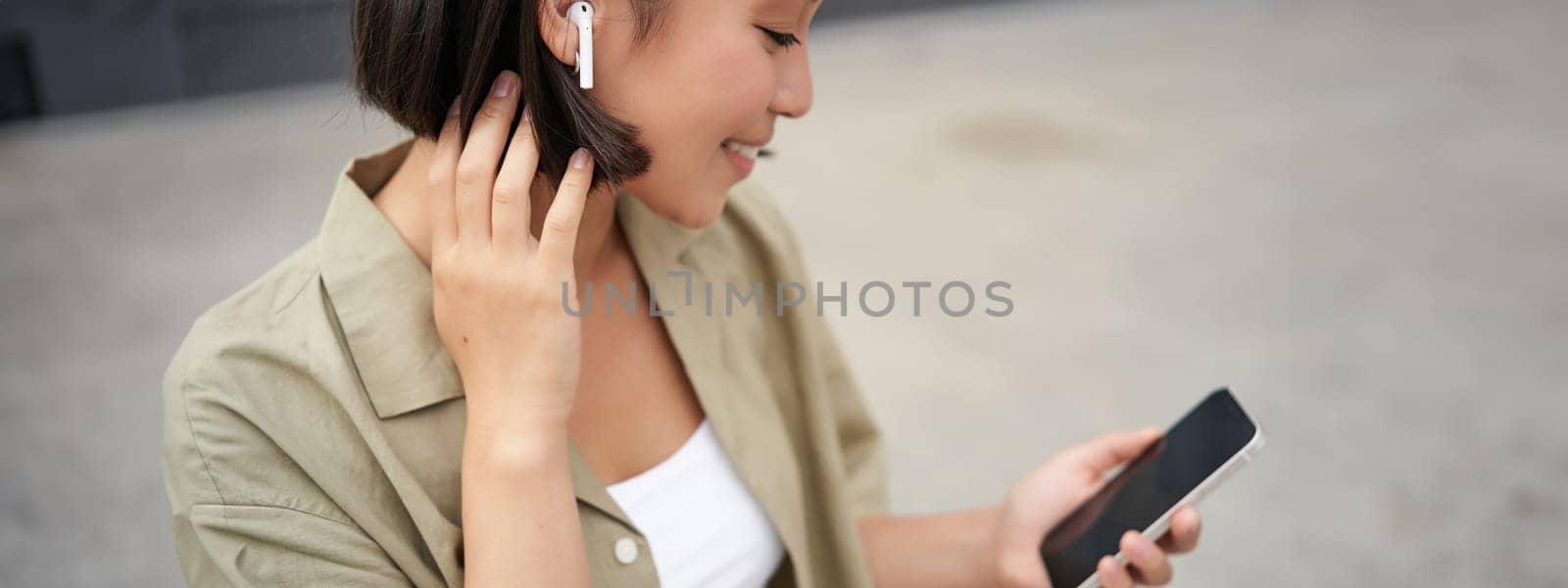 Close up portrait of asian girl, looking at mobile screen, listening music in headphones. Woman with earphones walks on street by Benzoix