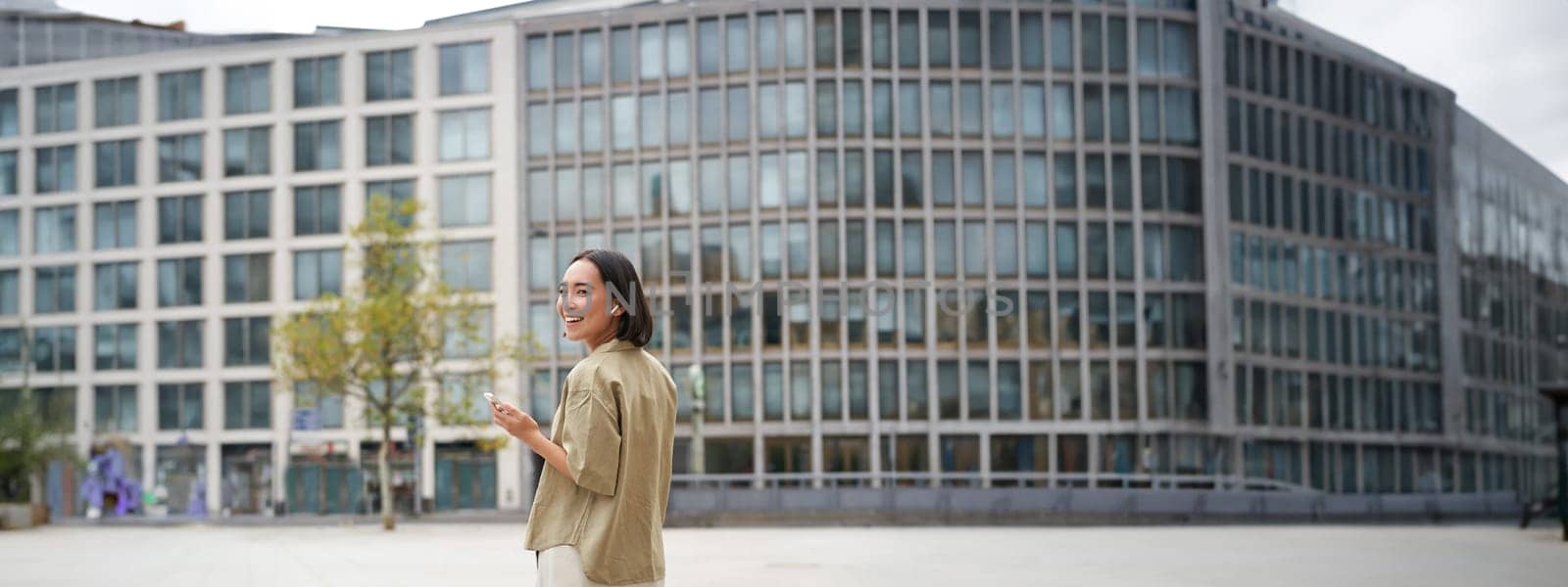 Silhouette of asian woman walking on street in wireless headphones, holding smartphone by Benzoix