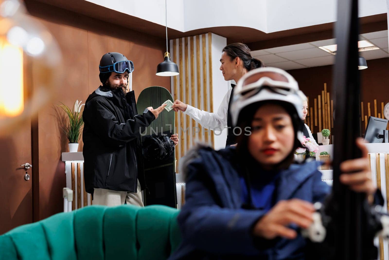 Image shows woman adjusting skiing equipment on sofa while man with snowboard gives money to concierge for good customer service. Male tourist tipping staff in hotel lobby as lady waits on couch.