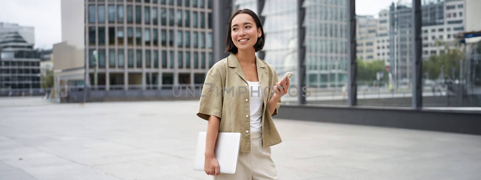 Asian girl with laptop and smartphone, standing on street of city centre, smiling at camera by Benzoix