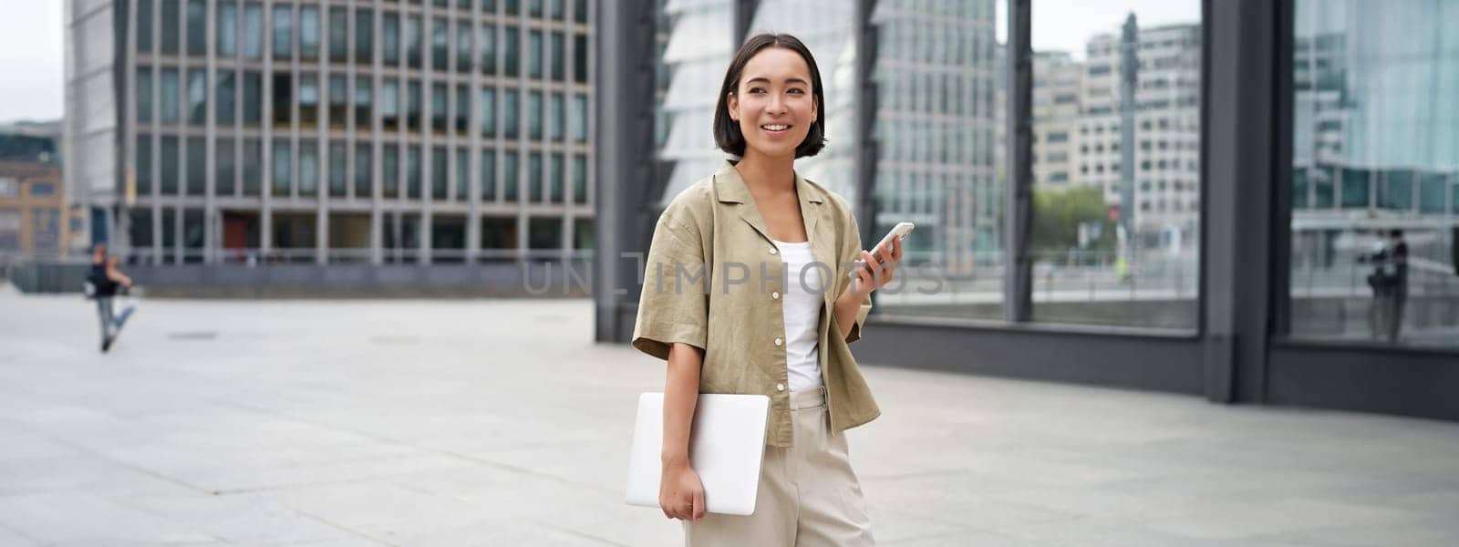 Asian girl with laptop and smartphone, standing on street of city centre, smiling at camera.