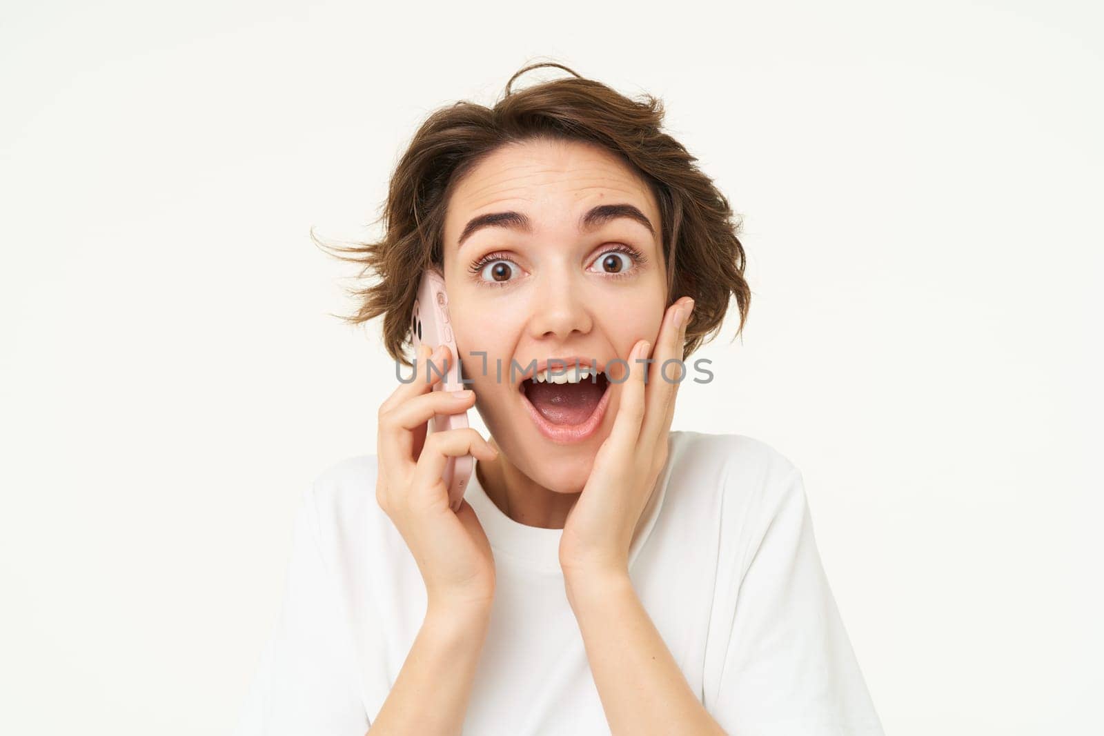 Portrait of woman with surprised face, answers phone call and looks excited, amazed by big news, stands over white background.