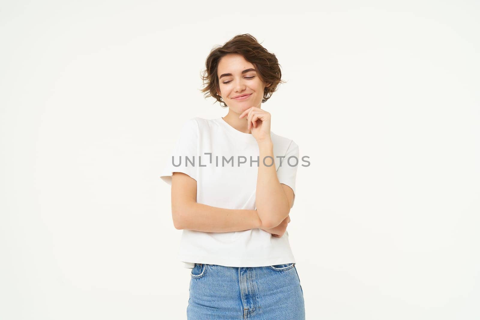 Image of pleased smiling brunette girl, smirks, looks down with confident, satisfied face expression, stands over white background.