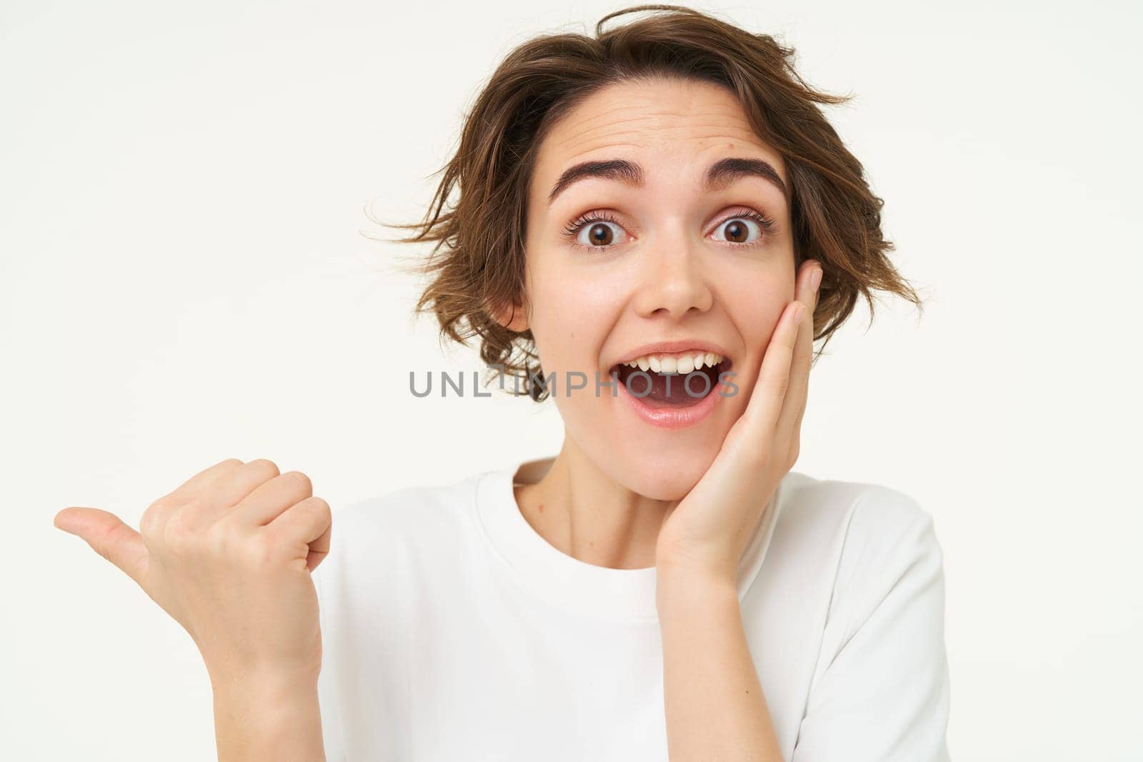 Image of girl with surprised face, points finger left, looks shocked, poses over white studio background. copy space