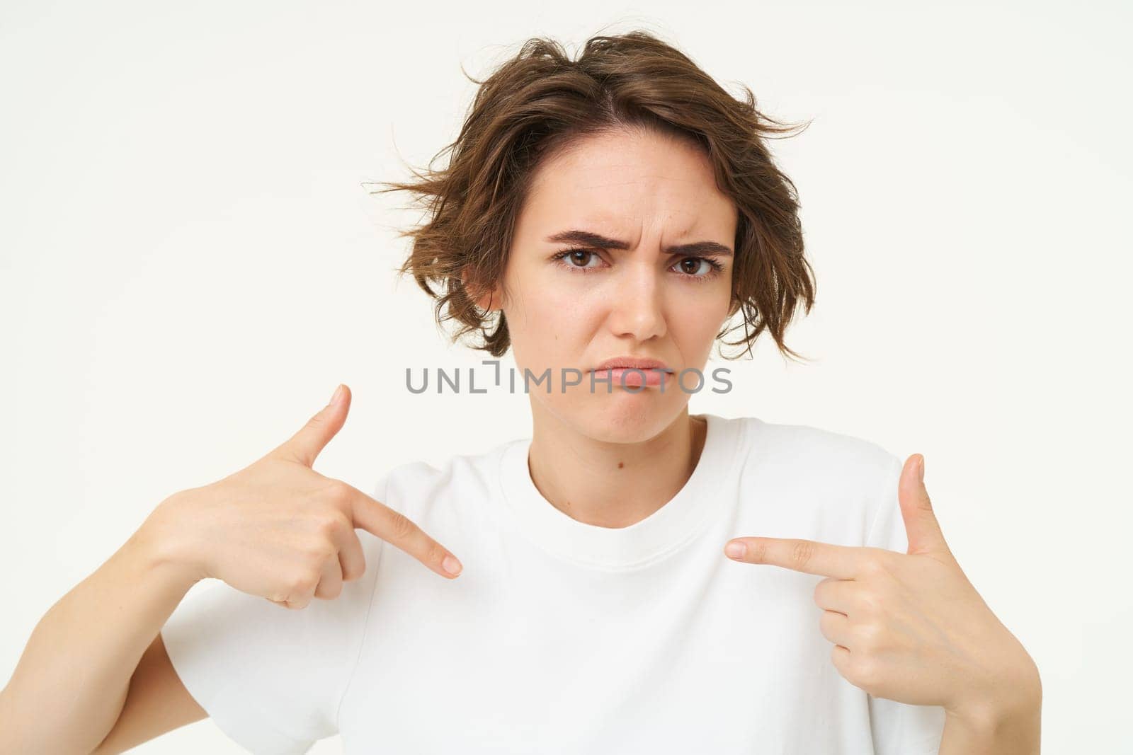 Close up portrait of woman with confused face, looks offended and points at herself, stands over white studio background.