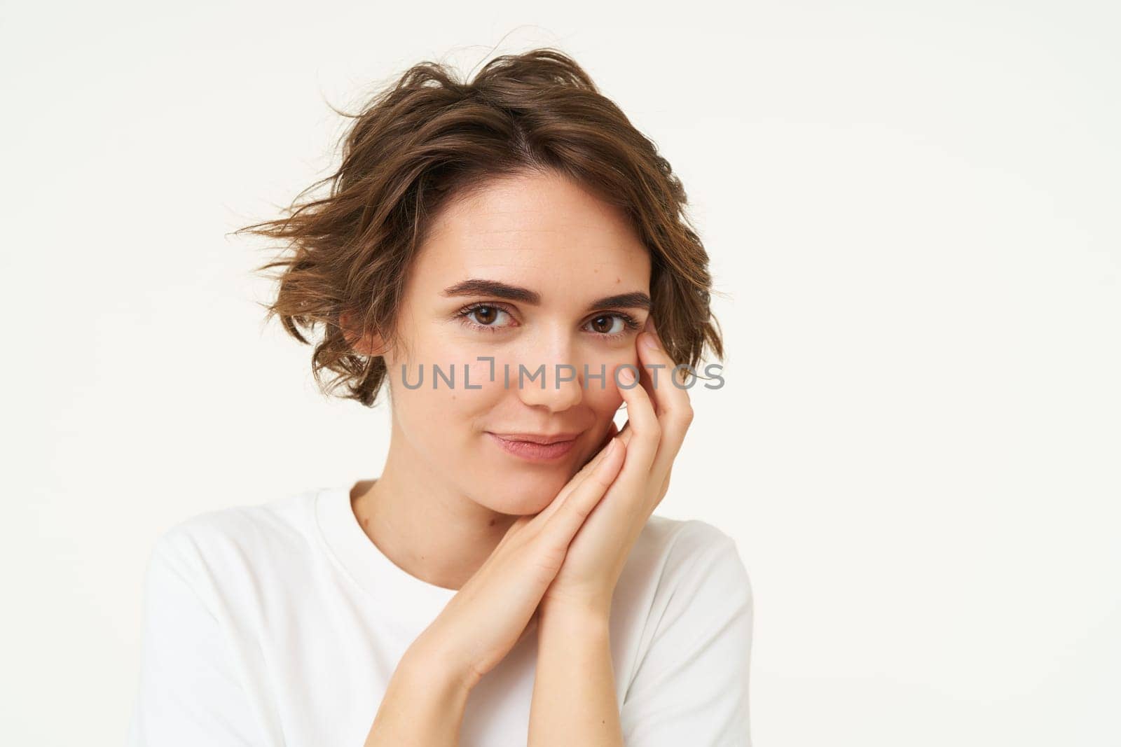 Close up of tender, feminine young woman, touches her face, looks gentle at camera, stands over white background.