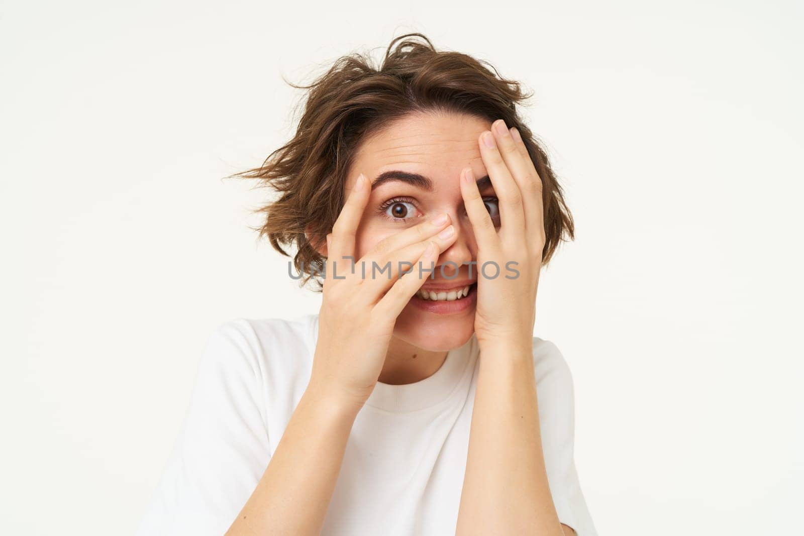 Image of funny woman peeking through fingers, looking at something with curious face expression, waiting for surprise and smiling excited, isolated over white background.