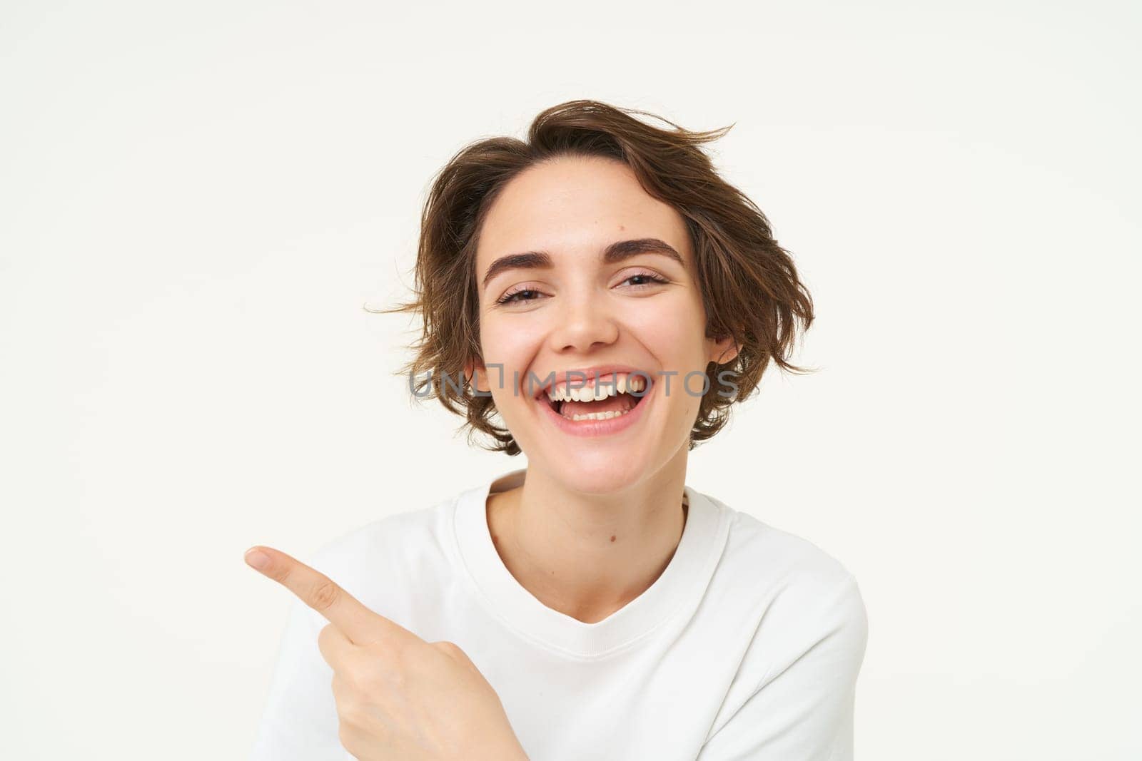 Close up shot of happy, candid woman, laughing and smiling, pointing finger left, showing advertisement, standing over white background by Benzoix