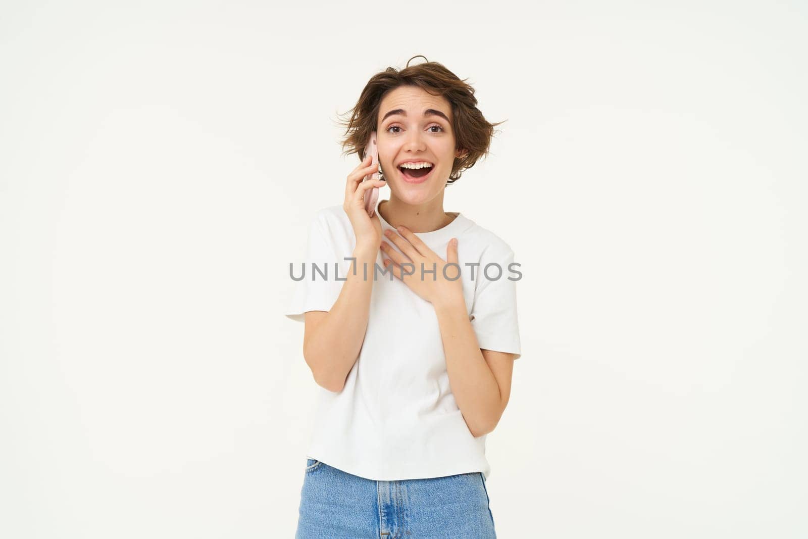 Portrait of chatty young woman talking on mobile phone, laughing and smiling, answer telephone with surprised face, standing over white background.