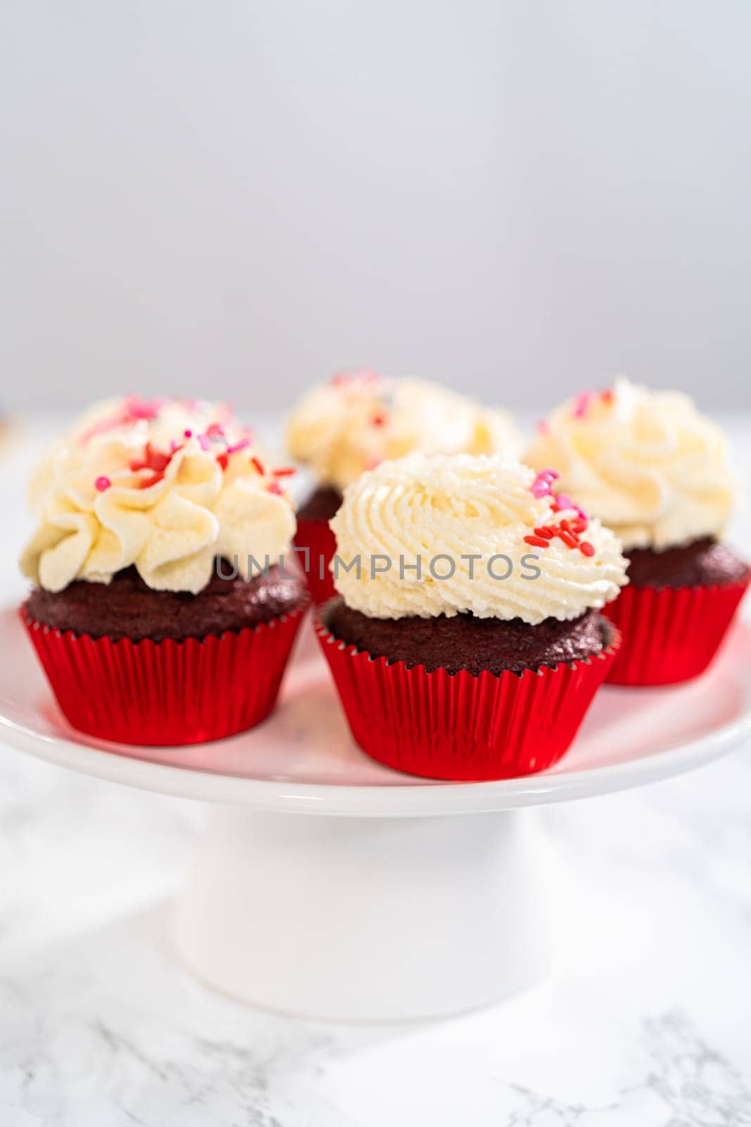 Freshly baked velvet cupcakes with white chocolate ganache frosting decorated with sprinkles.