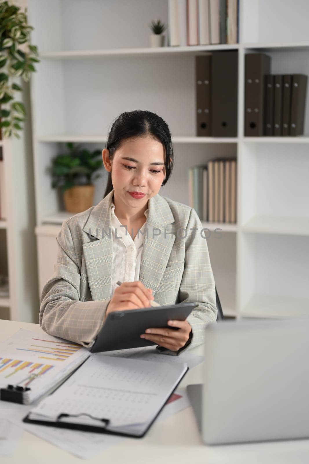 Attractive young businesswoman sitting at desk with documents and using a digital tablet.