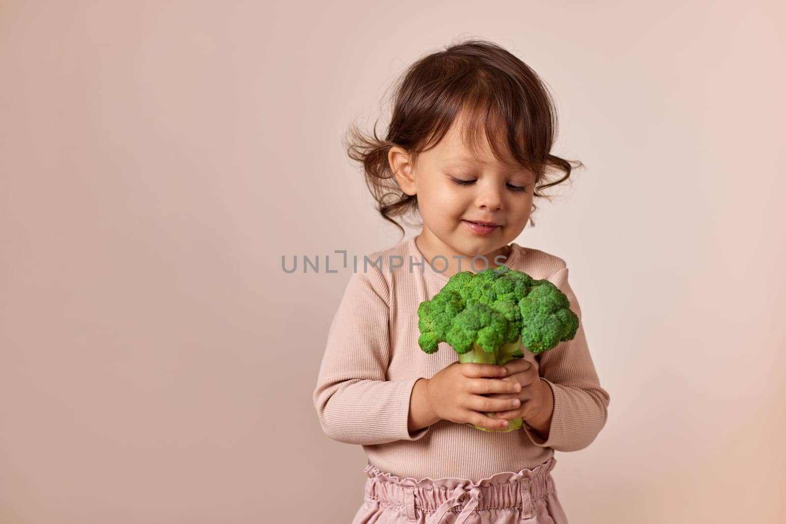 smiling child girl looking at fresh green raw broccoli on beige background. healthy baby food. kid loves vegetables