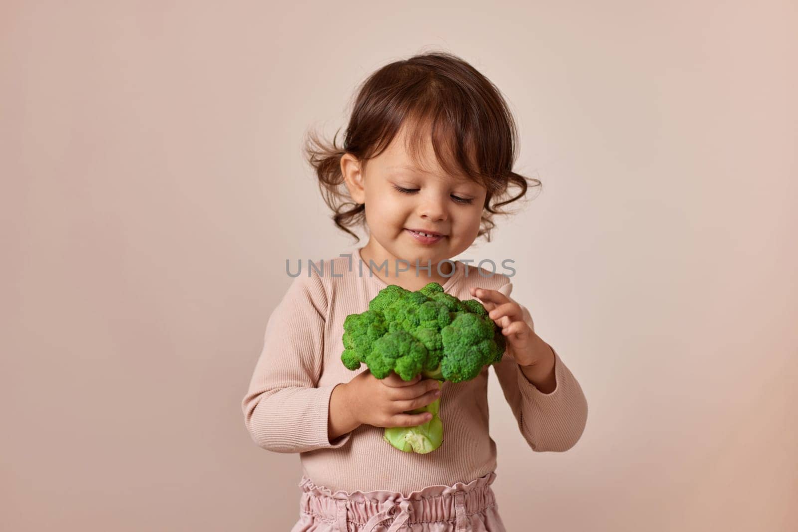 little child girl holding fresh green raw broccoli on beige background. healthy baby food. kid loves vegetables