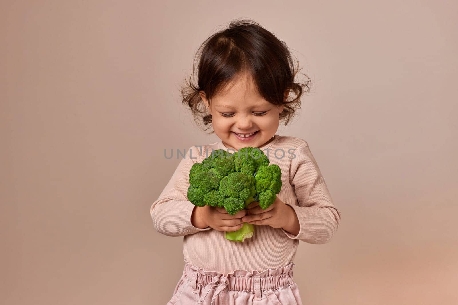 smiling cute child girl holding fresh green raw broccoli on beige background. healthy baby food. kid loves vegetables
