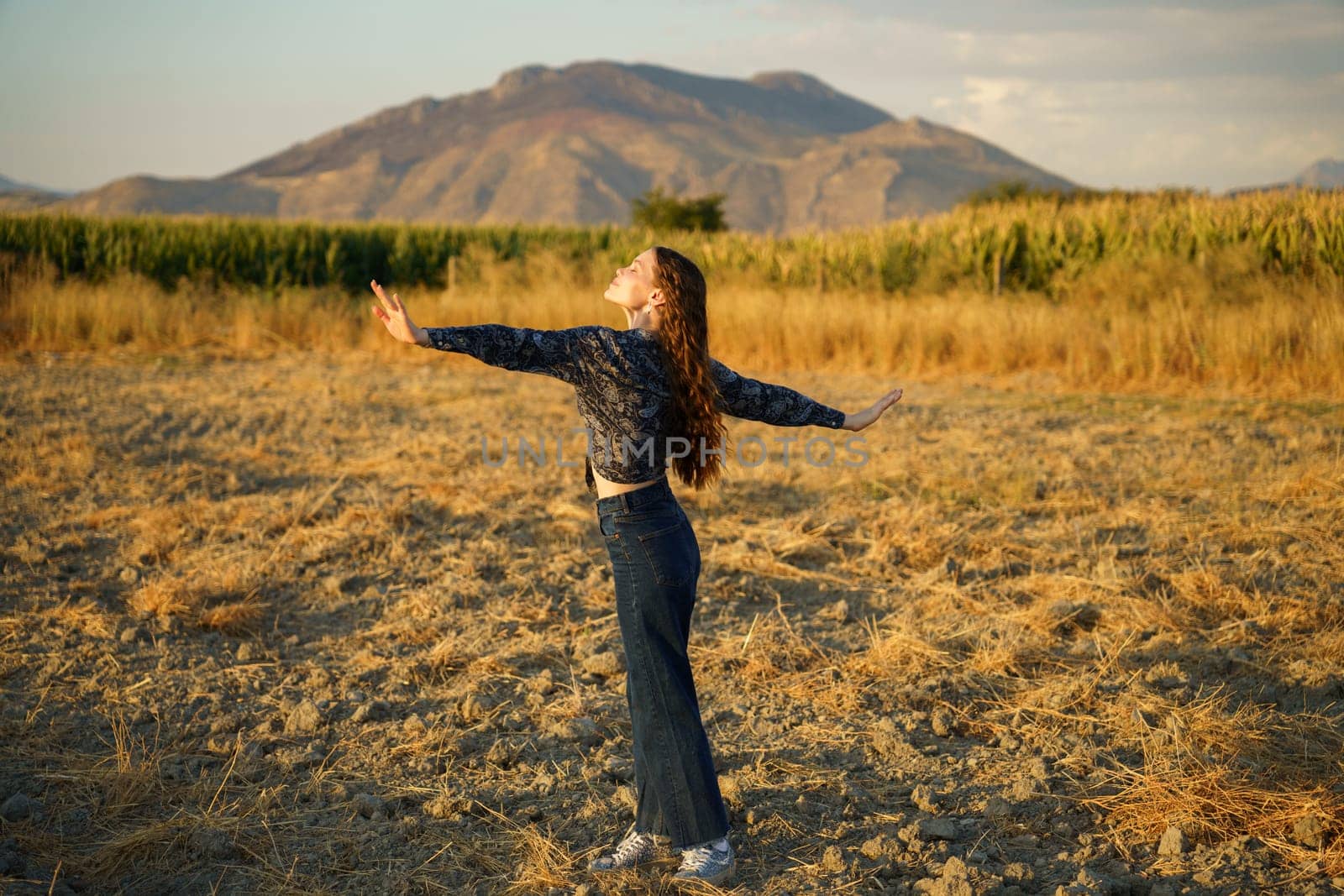 A carefree individual stands in the middle of a vast, open field, arms fully outstretched wide, delighting in the beauty and overwhelming tranquility of nature beneath a stunning mountain backdrop