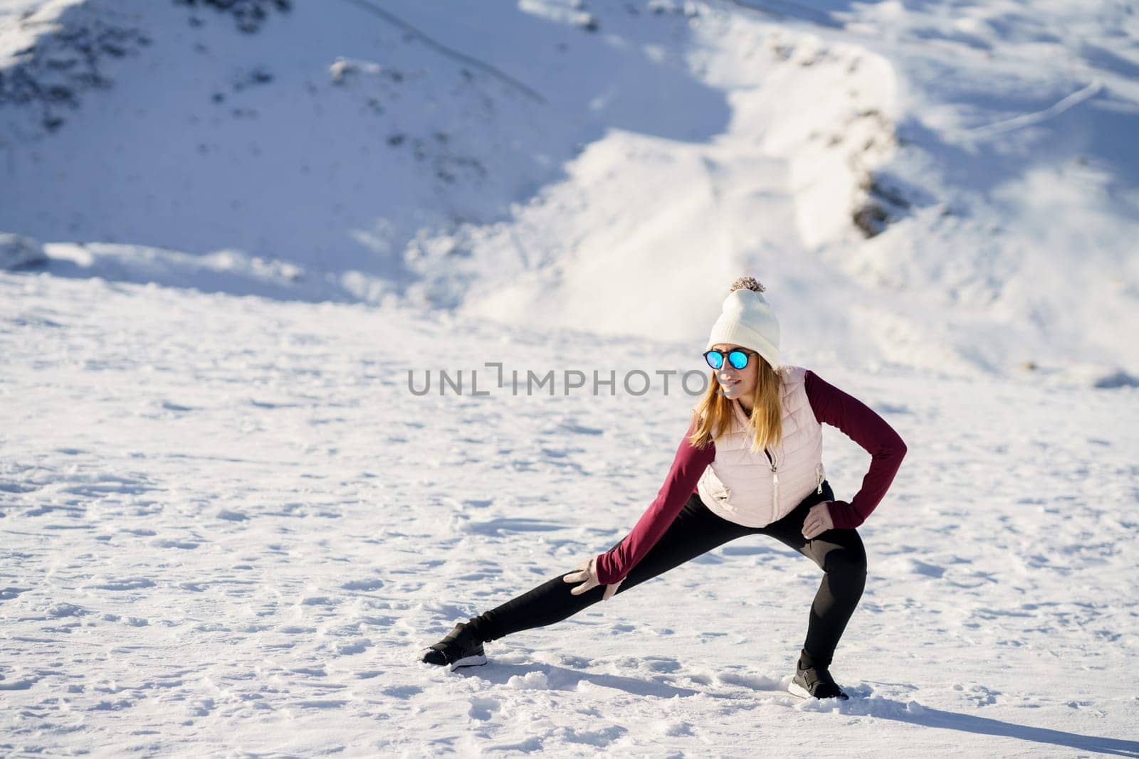 Full body of young female in warm clothes and sunglasses, standing on snowy slope in winter while practicing yoga with stretched legs and hands on knee warrior pose