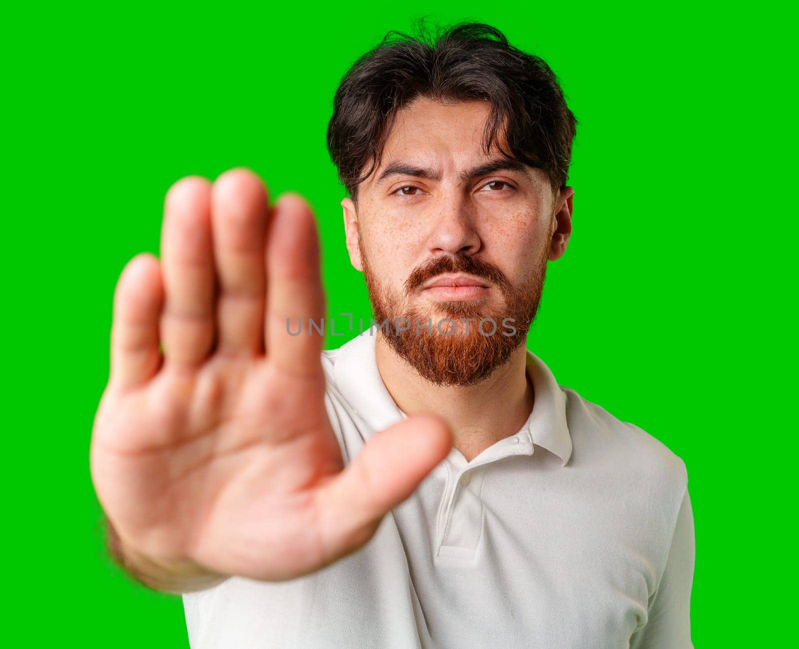 Man with a beard holding up his hand and looking at the camera against a bright green background in a neutral setting