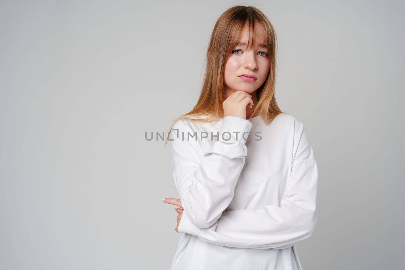Young woman in a white long-sleeve shirt thoughtfully poses with a hand on her chin in a minimalistic indoor setting