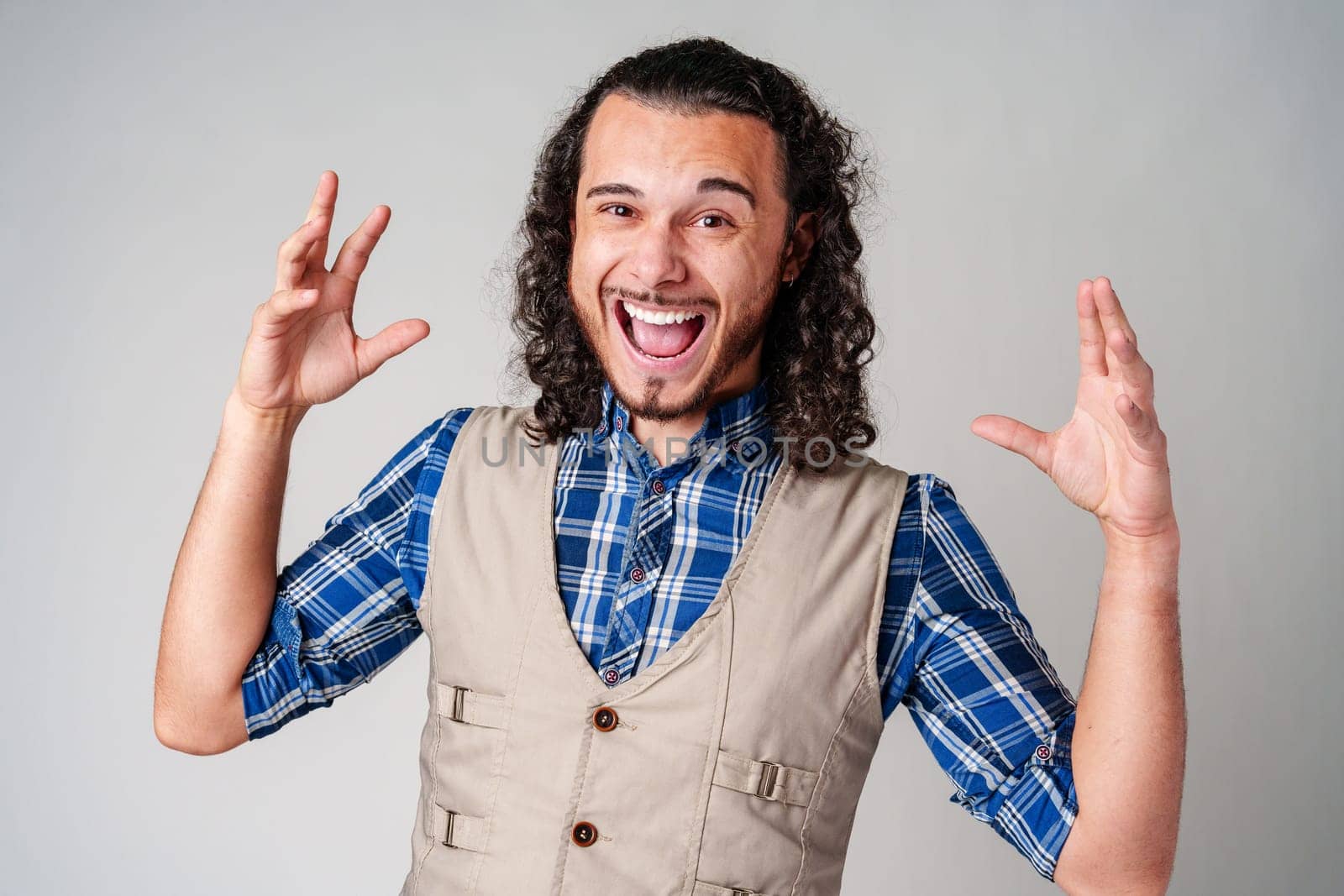 A man with curly hair excitedly gesturing while wearing a plaid shirt and vest against a plain background close up