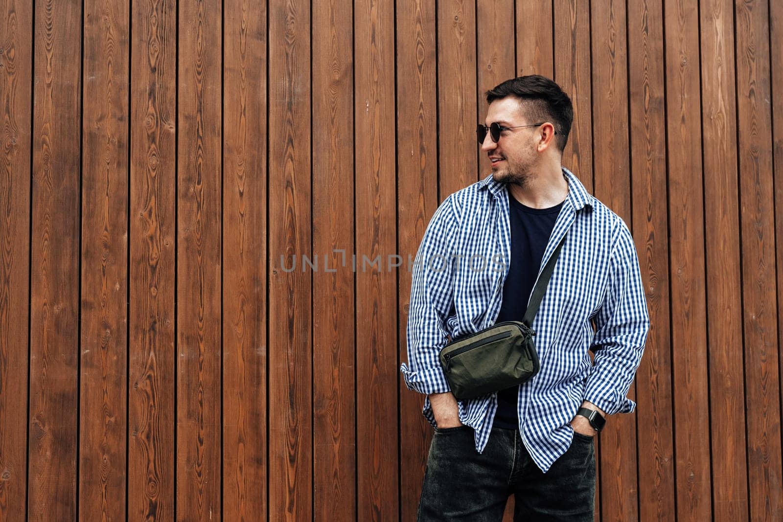 A man in a blue checkered shirt smiles while standing outside a modern building on a sunny day close up