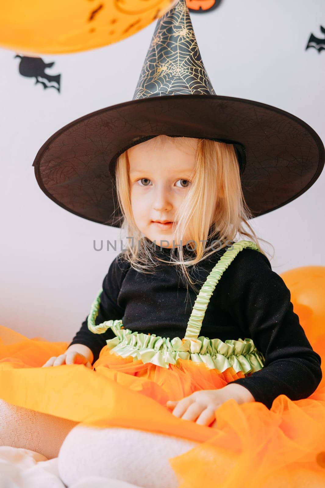 Children's Halloween - a girl in a witch hat and a carnival costume with airy orange and black balloons at home. Ready to celebrate Halloween.