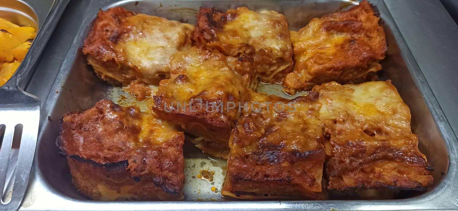 A generous serving of homemade lasagna is presented in a metal tray, showcasing layers of pasta, cheese, and sauce, enticing guests at a family gathering in the evening.