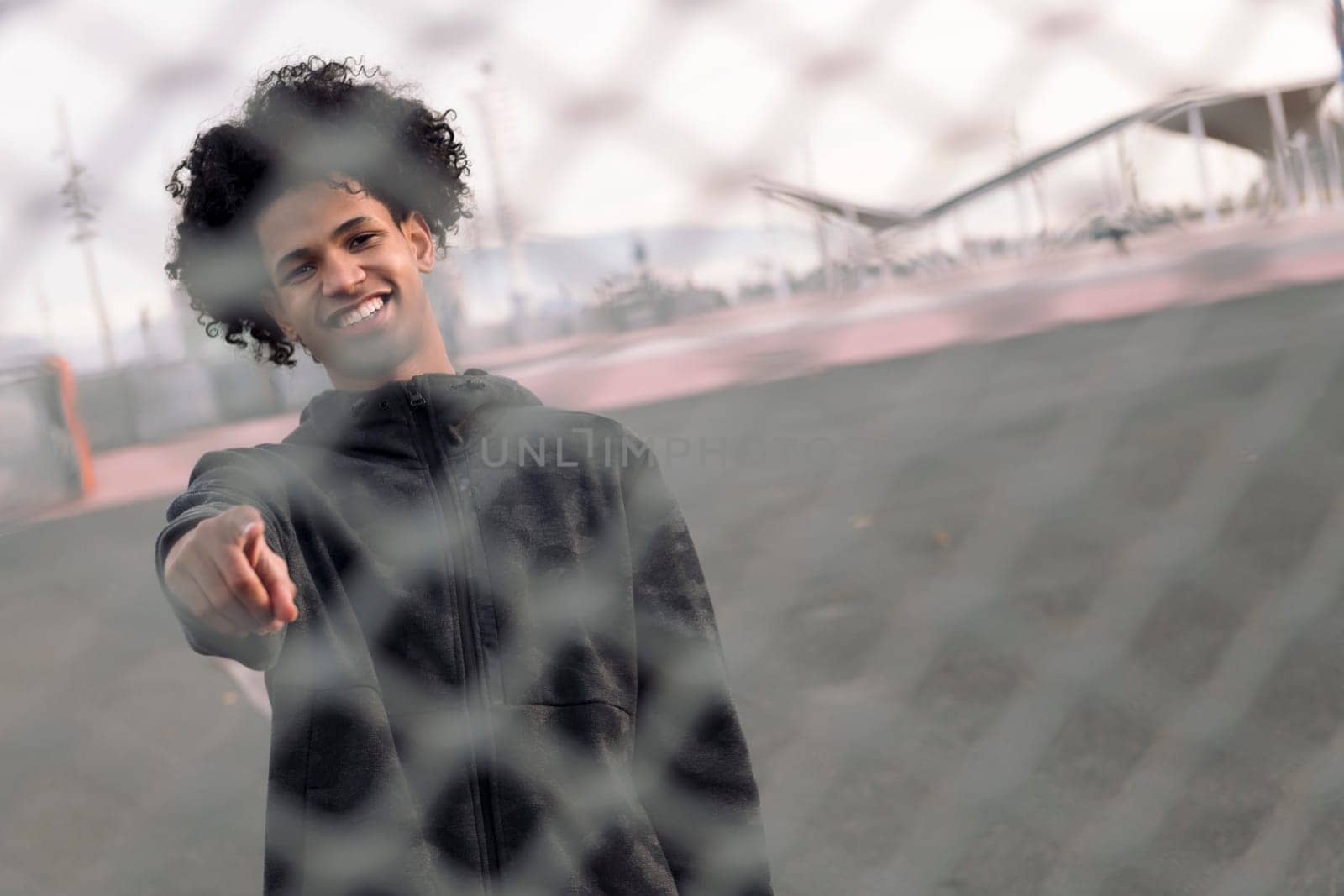 smiling teen african american male with curly hair pointing looking at camera while standing behind metal chain link fence on urban street, concept of adolescence and young lifestyle