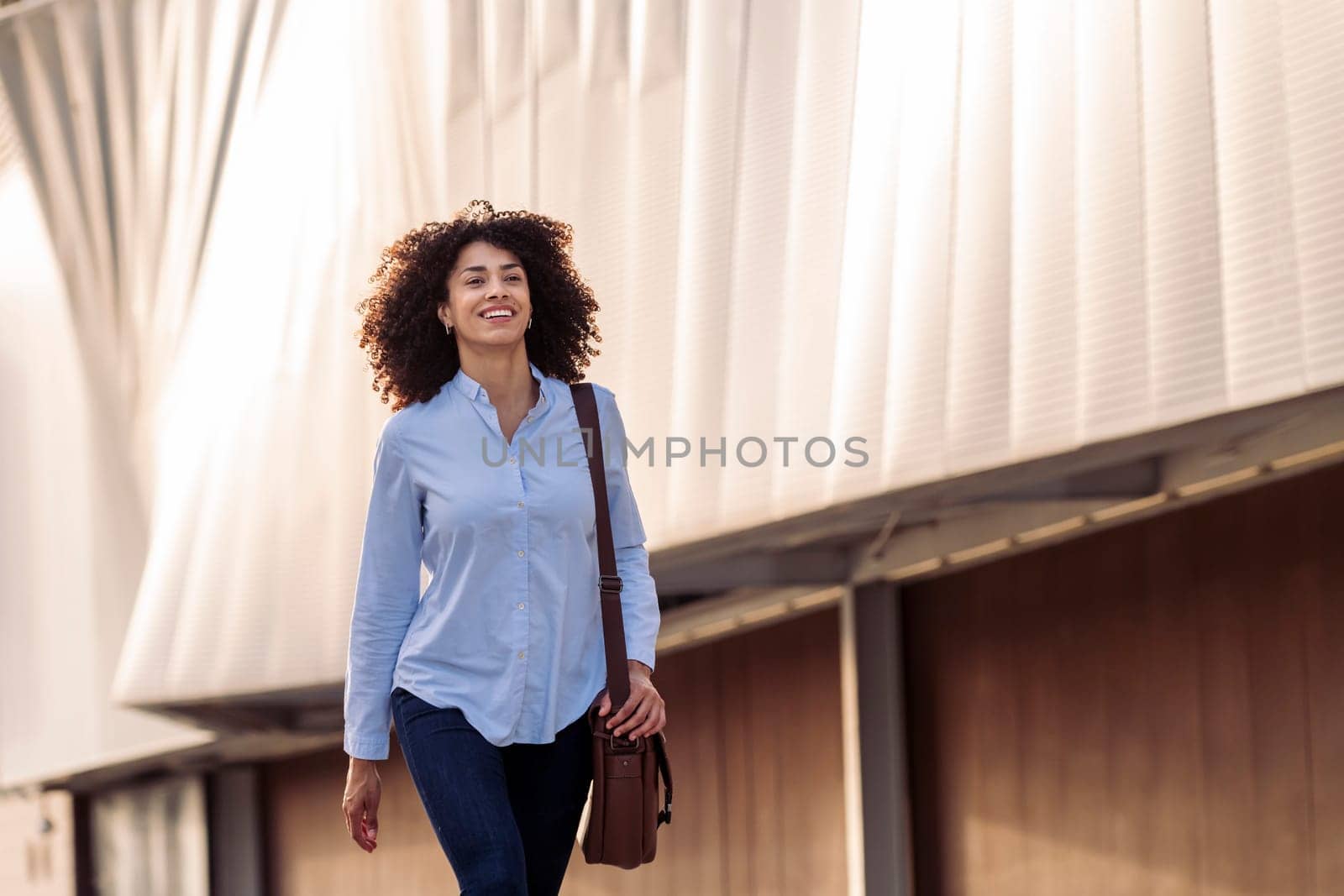 satisfied ethnic businesswoman with bag walking along street and smiling while looking forward