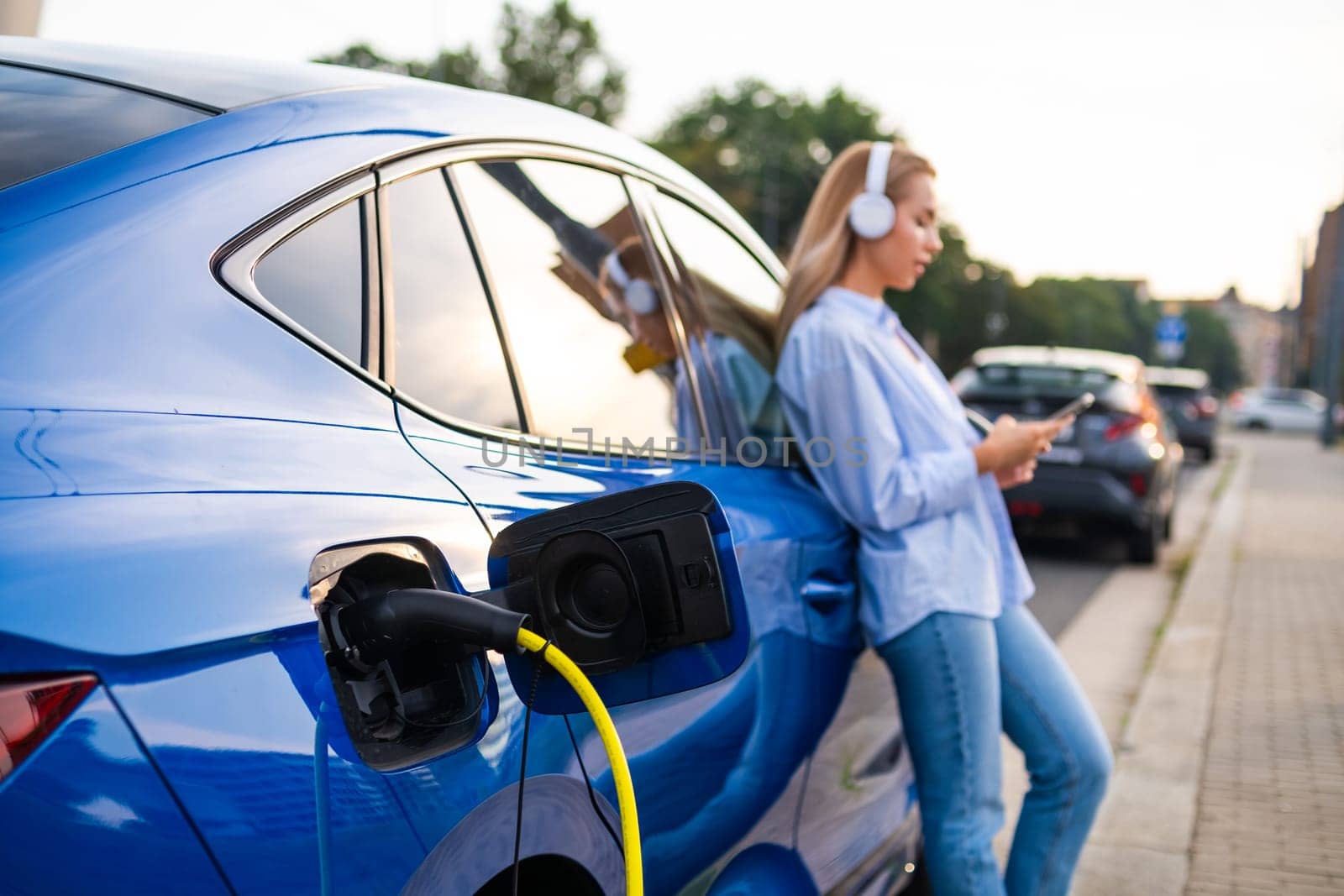 Close up electric car charging by the roadside, while a woman stands nearby wearing headphones, listening to music