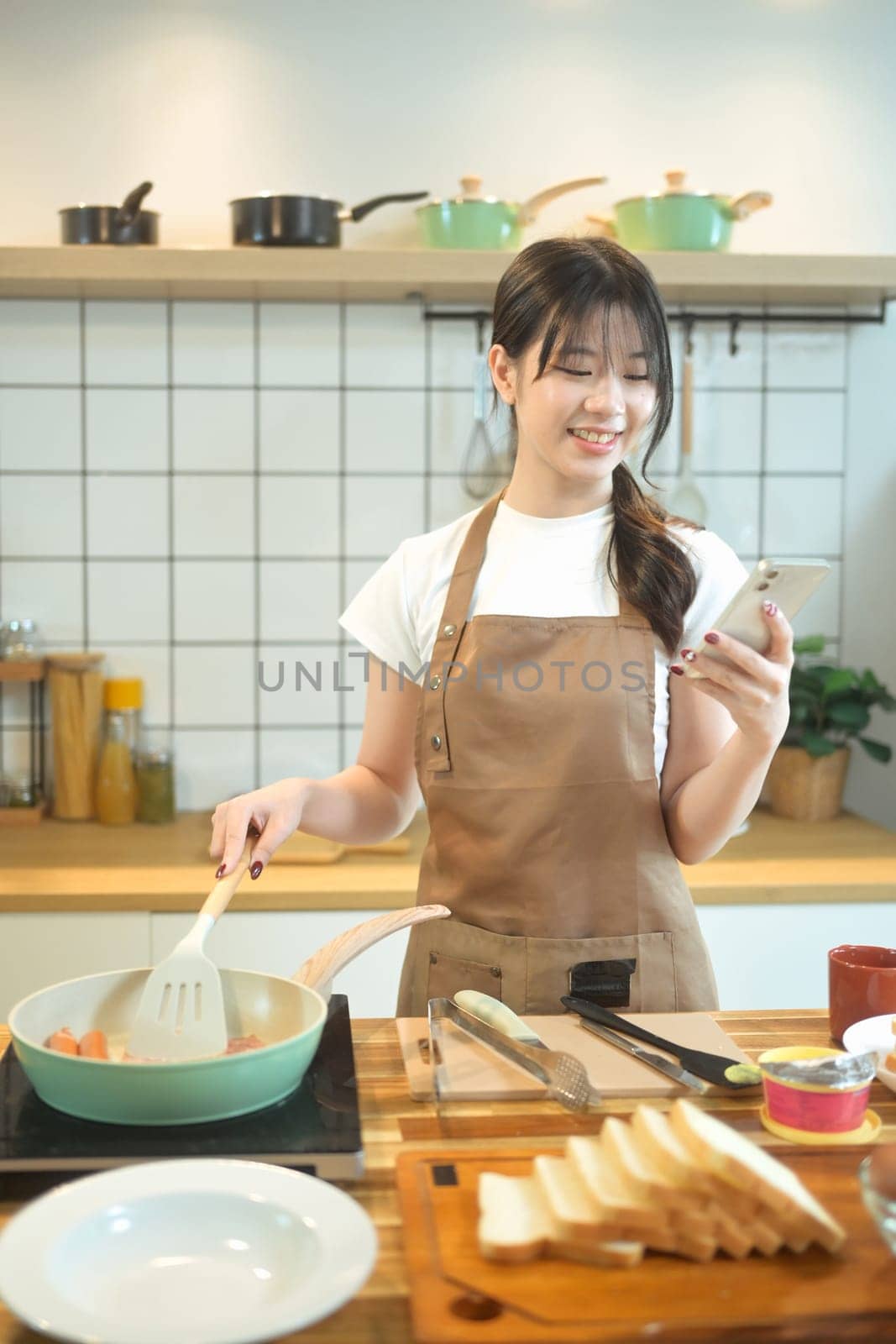 Smiling young woman using smartphone while preparing meal on a wooden worktop.
