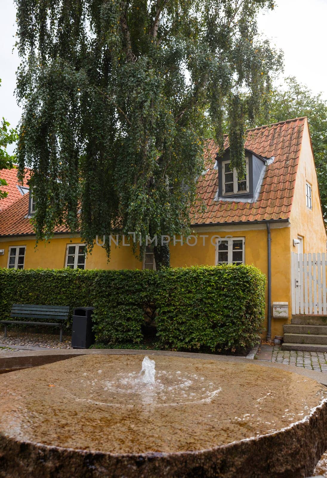 ebeltoft,denmark,A quaint yellow house with a red-tiled roof stands amidst lush greenery. A rustic stone fountain in the foreground adds a tranquil, serene atmosphere to the picturesque scene