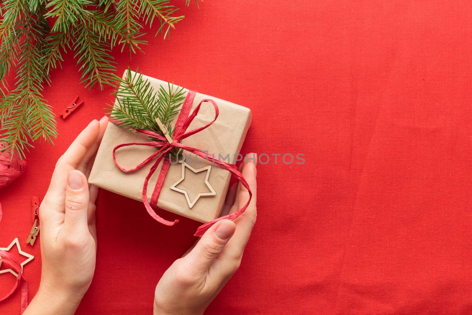 Female hands holding Merry Christmas eco-friendly natural packaging gift box on red decorated festive background, woman prepare a surprise for the new year, winter handmade holiday green present concept, flat lay, top view