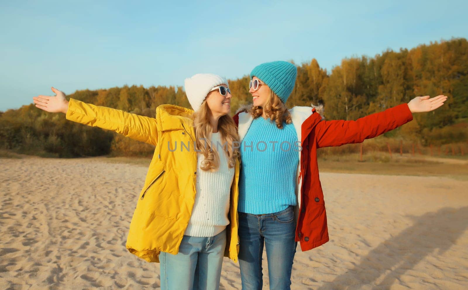 Happy joyful young two women friends together on sunny beach, stylish smiling girlfriends standing on sea coast in glasses, colorful red yellow coat jacket and hat outdoors