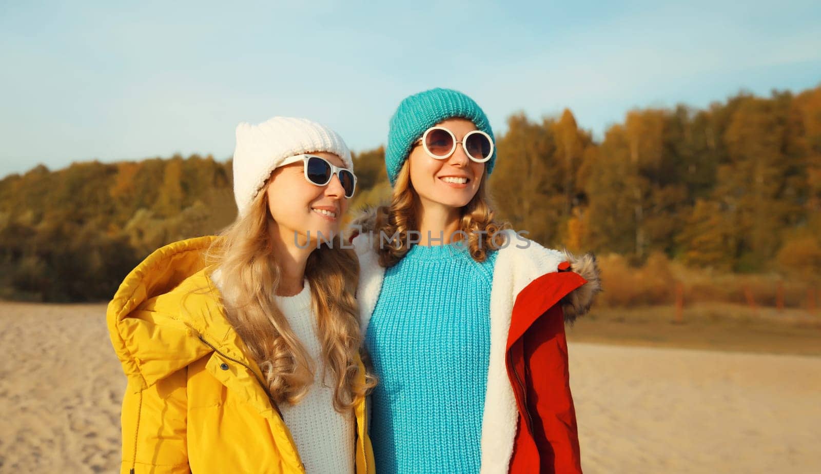 Happy young two women friends together on sunny beach, stylish smiling girlfriends looking away standing on sea coast in glasses, colorful red yellow coat jacket and hat outdoors