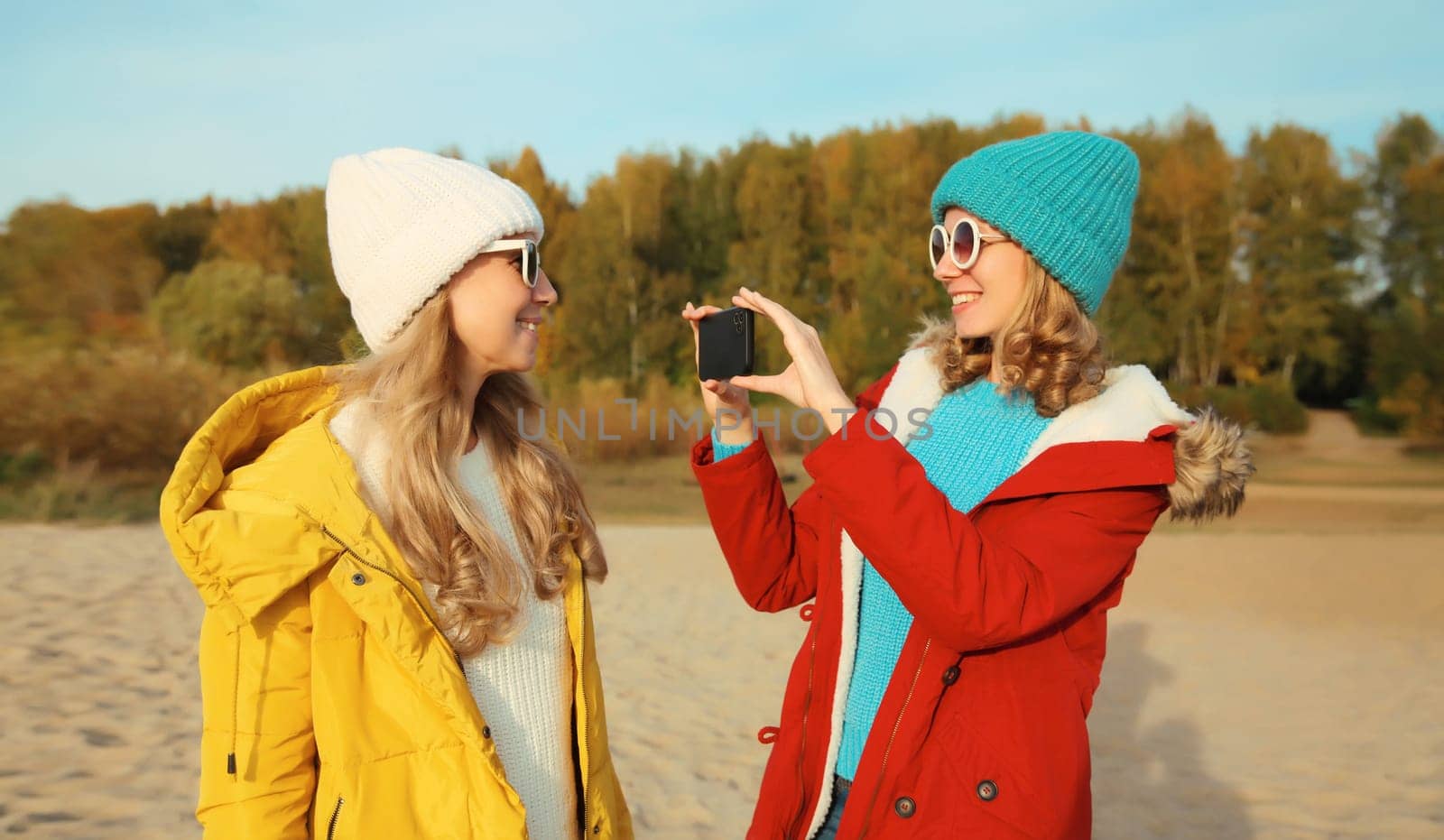Happy joyful young two women friends taking selfie with smartphone together on sunny beach, stylish smiling girlfriends having fun on sea coast in glasses, jacket, hat outdoors