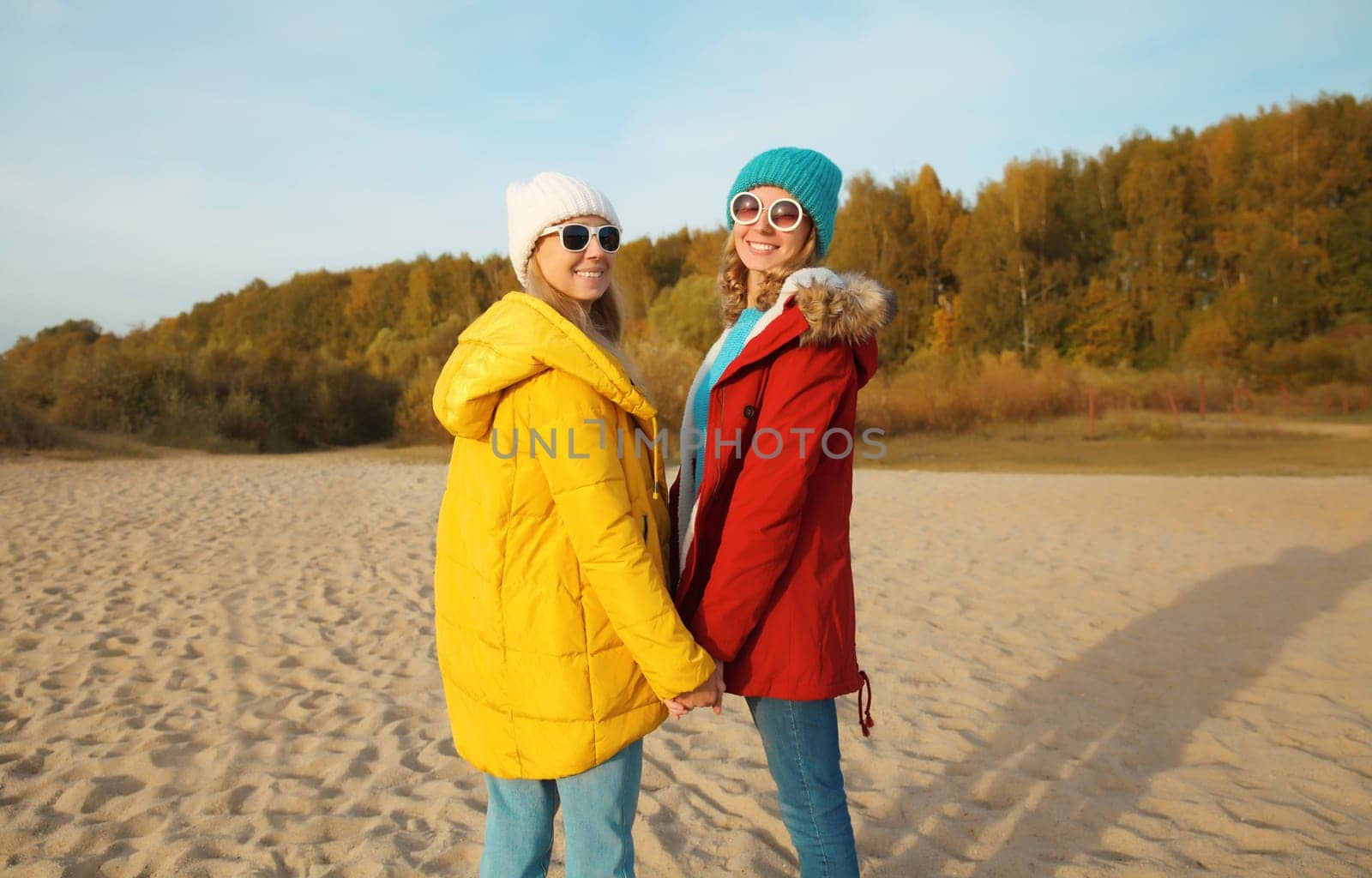 Happy young two women friends together on sunny beach, stylish smiling girlfriends standing on sea coast in glasses, colorful red yellow coat jacket and hat outdoors