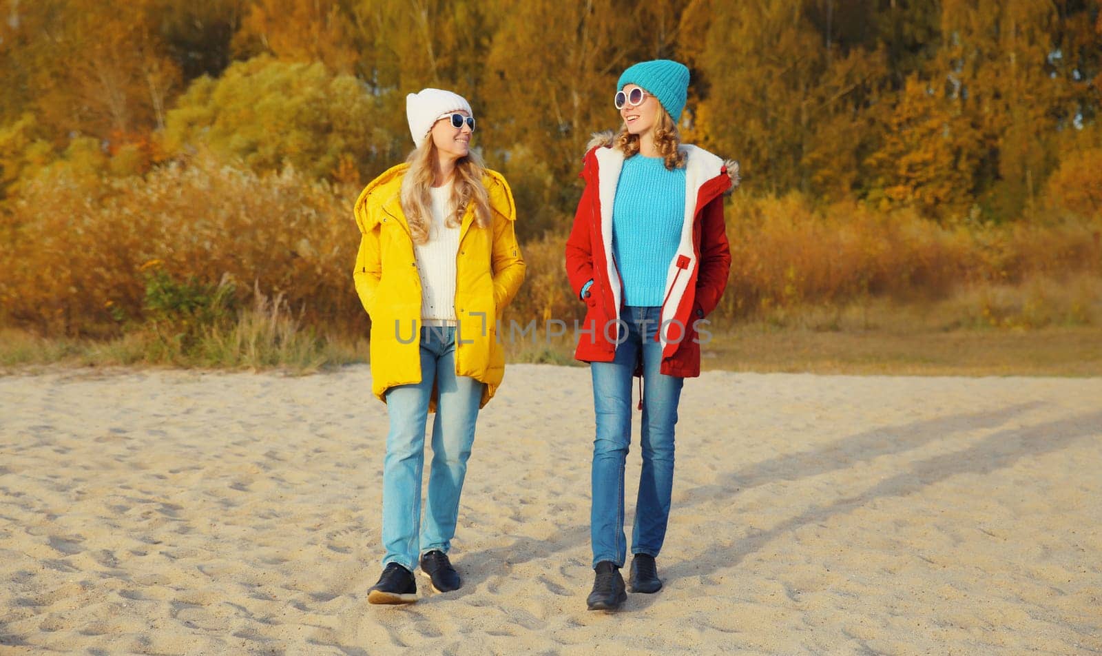 Happy young two women friends walking and talking together on sunny beach, stylish smiling girlfriends on sea coast in glasses, colorful red yellow coat jacket and hat outdoors on a warm day