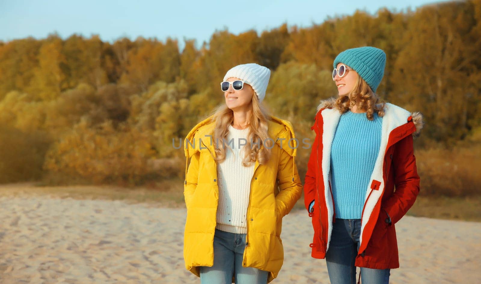 Happy young two women friends together on sunny beach, stylish smiling girlfriends looking away standing on sea coast in glasses, colorful red yellow coat jacket and hat outdoors