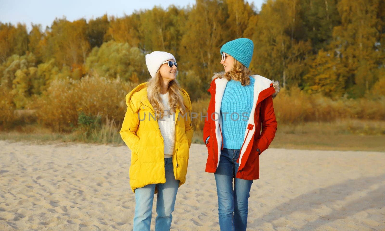 Happy young two women friends walking and talking together on sunny beach, stylish smiling girlfriends on sea coast in glasses, colorful red yellow coat jacket and hat outdoors on a warm day